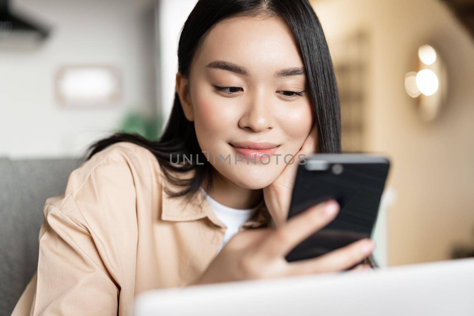 Portrait of asian girl sitting with laptop, checking her phone and smiling, browsing websites on smartphone by Benzoix
