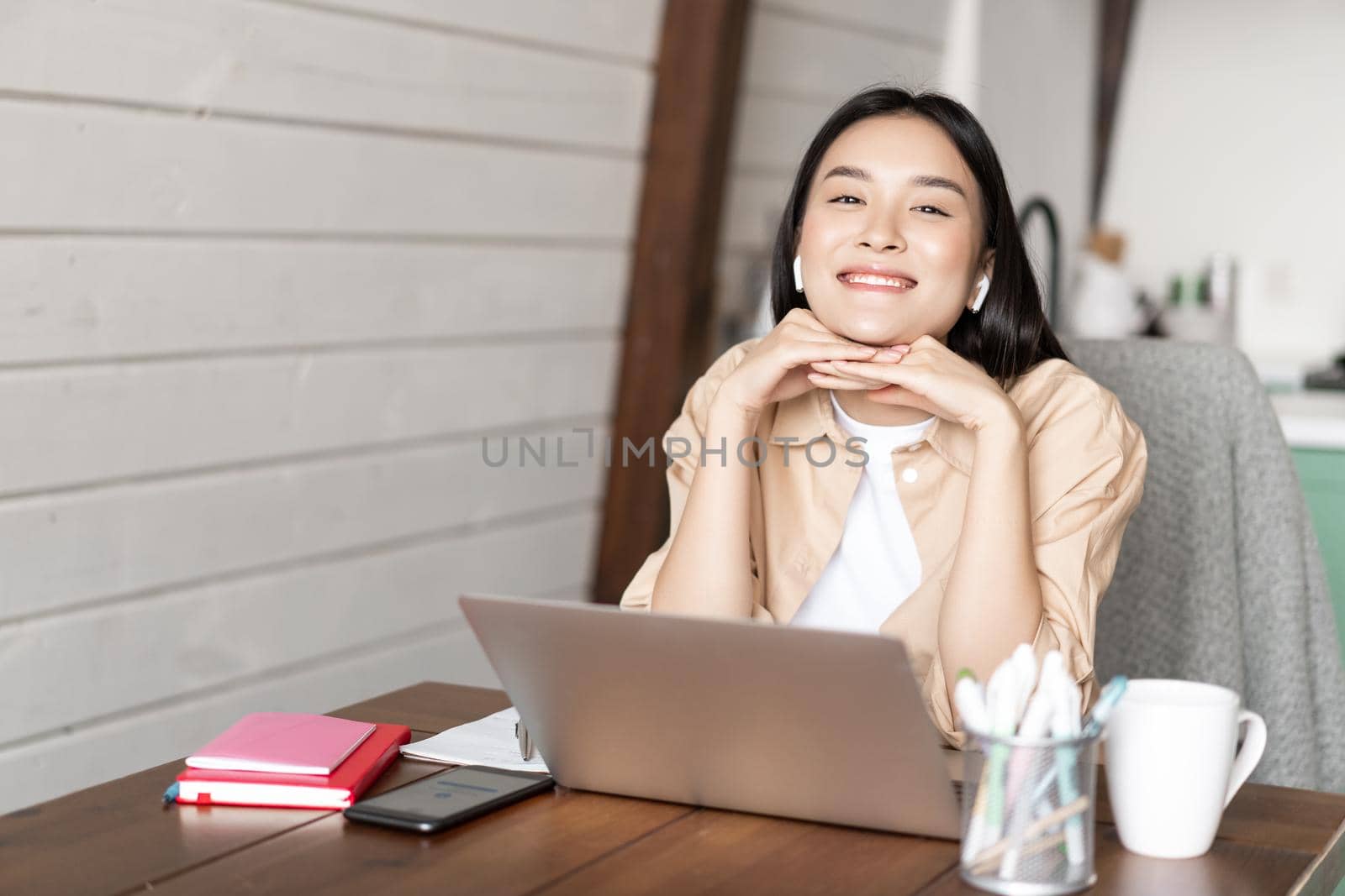 Online education and work from home concept. Smiling asian girl sits with laptop and earphones in kitchen, working remotely, having conference or video chat.