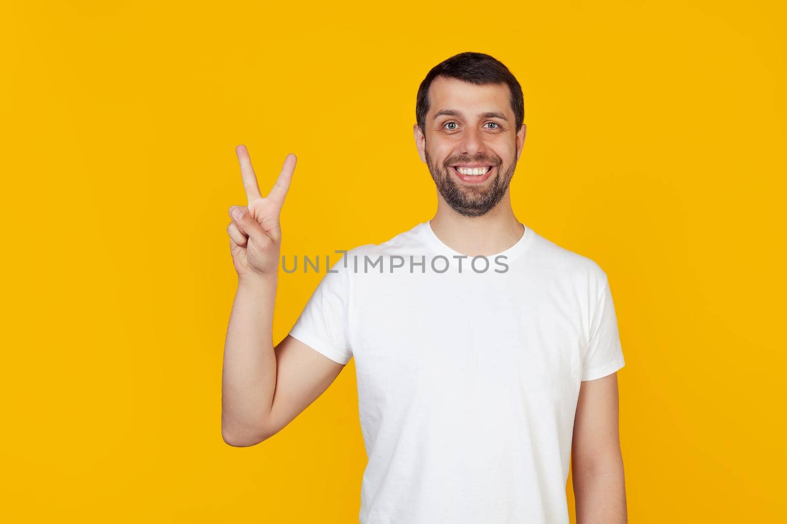 Modern young man with a beard in a white tank top shows number two with fingers on hand, smiling confidently and happily, looking into the camera The man shows two fingers. Number 2 yellow background.