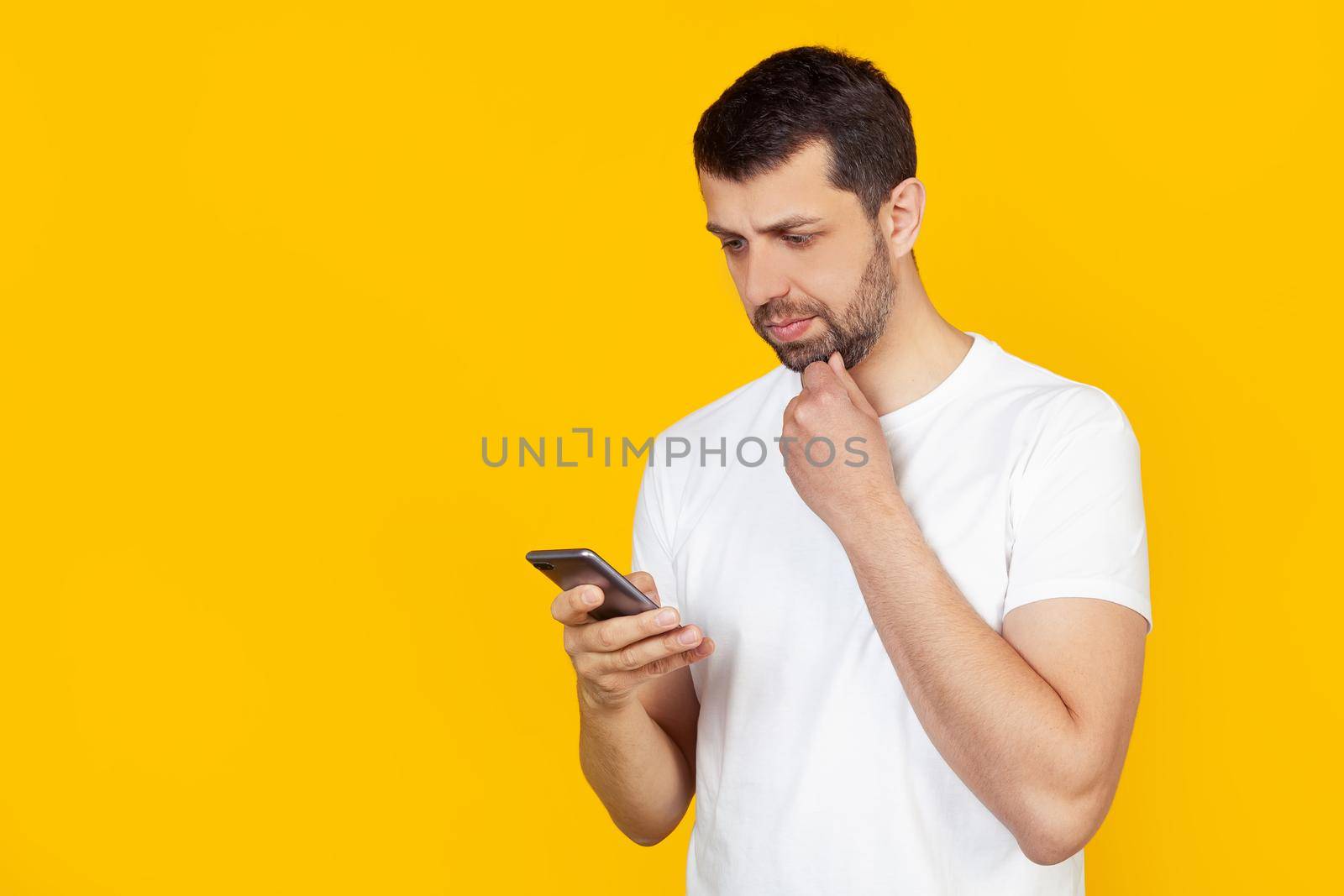 A young man with a beard in a white t-shirt uses a smartphone with a confident expression on his smart face, thinking seriously. Stands on isolated yellow background.