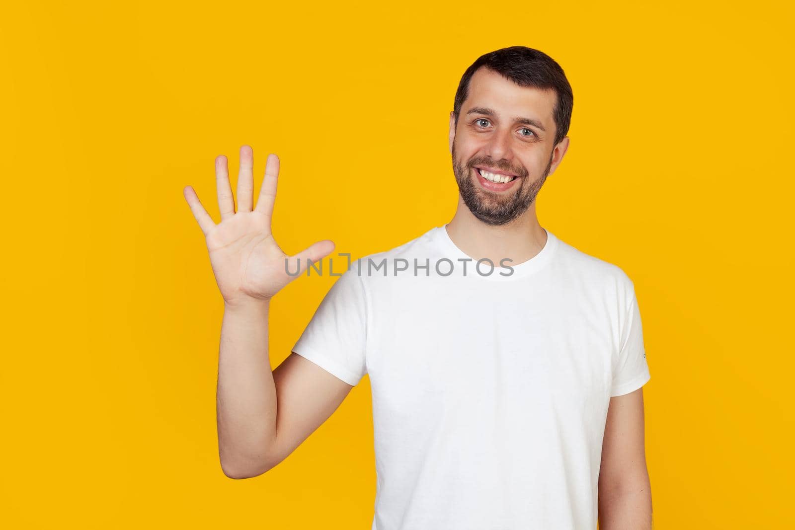 Modern young man with a beard in a white tank top shows number five with fingers on hand, smiling confidently and happily, looking at the camera. The man shows five fingers. Number 5 yellow background
