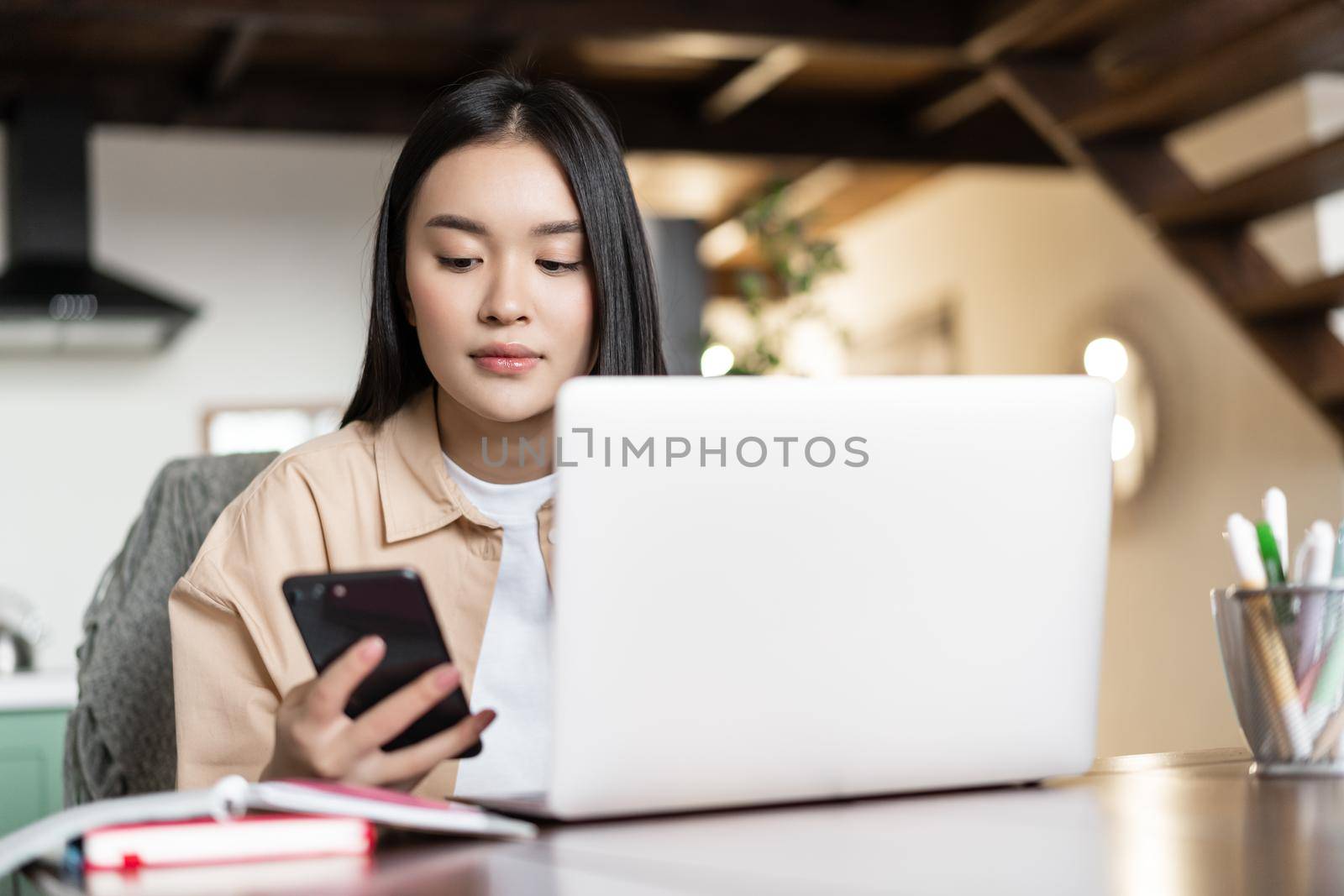 Remote work and distance education concept. Young asian business woman working with laptop, checking mobile phone, doing homework on computer at home.