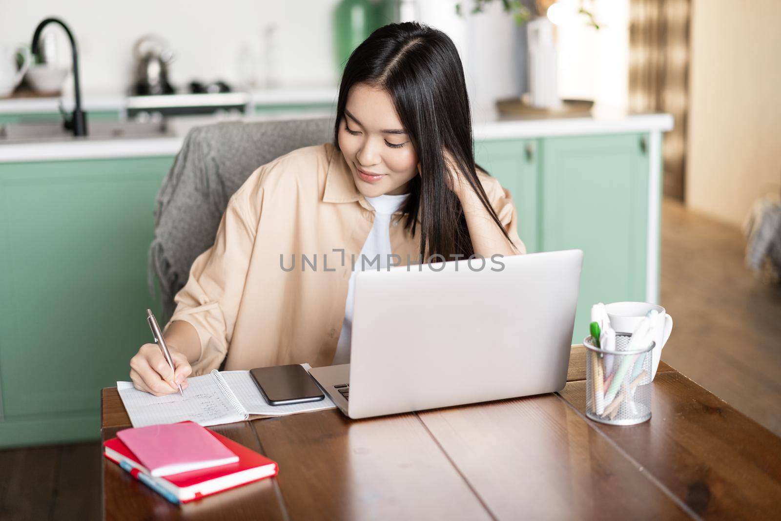Asian girl student doing homework at home. Young korean woman writing with pen, using laptop to study online, working from indoors.