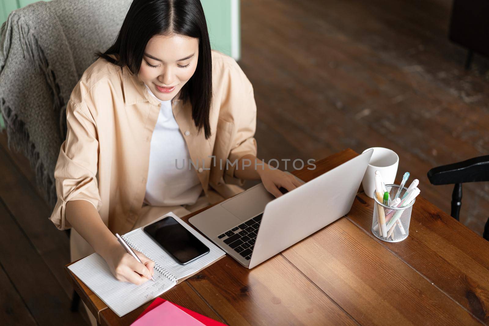 Upper angle of asian girl working indoors. Woman studying remotely, writing down notes, doing homework with laptop and smartphone.