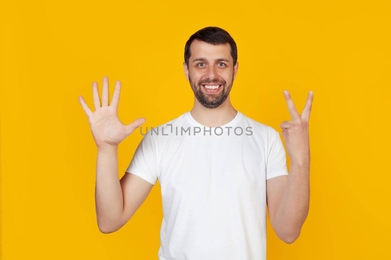 Modern young man with a beard in a white tank top shows number seven with fingers on hand, smiling confidently and happily, looking into the camera. The man shows seven fingers. Number 7.