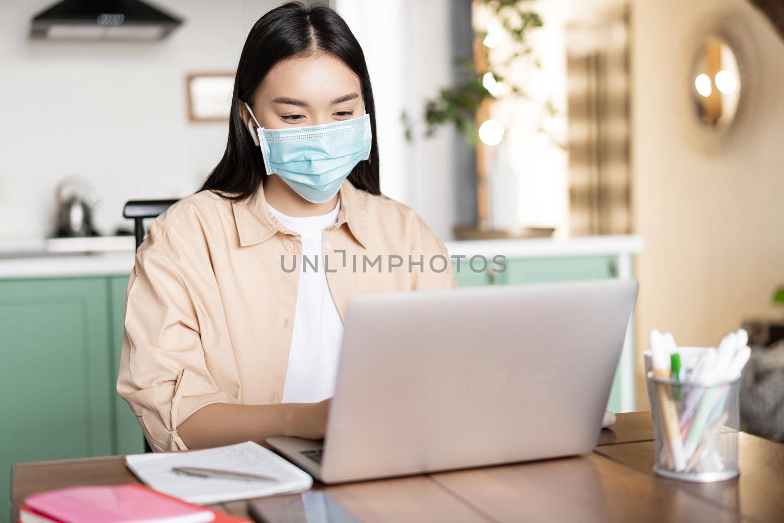 Asian girl in medical mask working from home while being sick with covid, using laptop by Benzoix