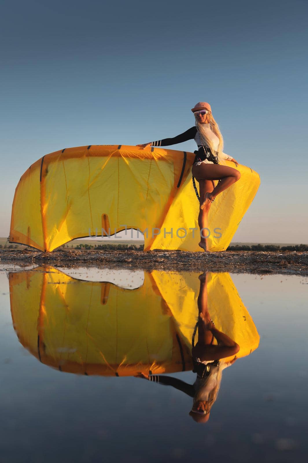 Attractive stylish young caucasian woman in cap sunglasses and kitesurfer outfit standing next to sandy shore next to her kite at sunset reflecting in the water.