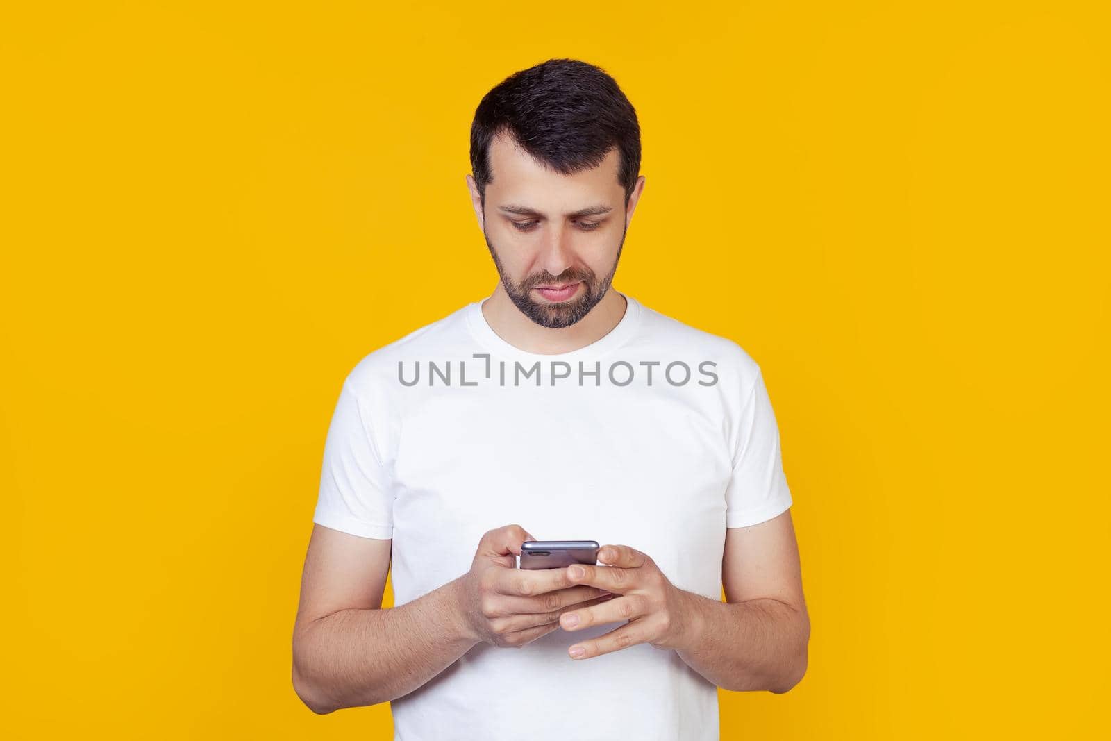 A young man with a beard in a white T-shirt holds in his hand and looking into a smartphone looking for information on the Internet, a natural expression, stands on an isolated yellow background.