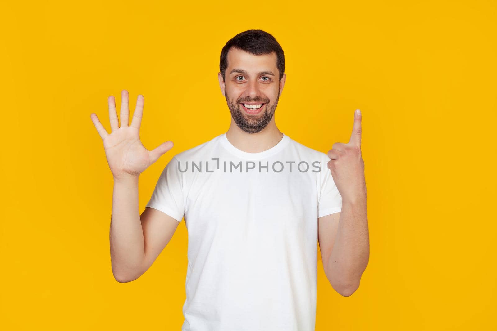 Modern young man with a beard in a white tank top shows number six with fingers on hand, smiling confidently and happily, looking into the camera The man shows six fingers. Number 6. yellow background