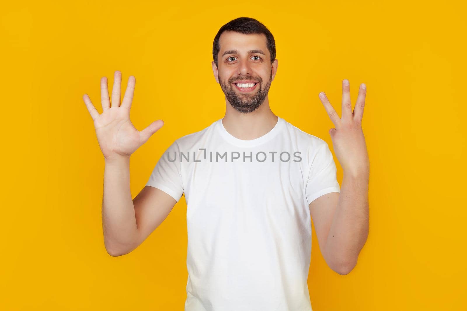 Modern young man with a beard in a white tank top shows number eight with fingers on hand, smiling confidently and happily, looking into the camera. The man shows eight fingers. Number 8.