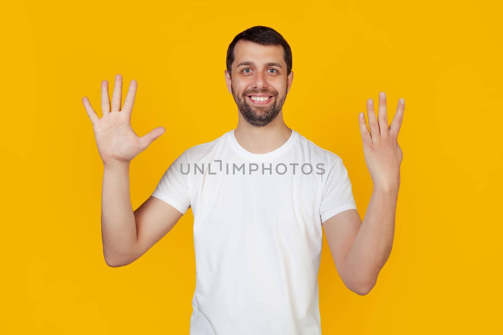 Modern young man with a beard in a white tank top shows number nine with fingers on hand, smiling confidently and happily looking into the camera The man shows nine fingers. Number 9 yellow background