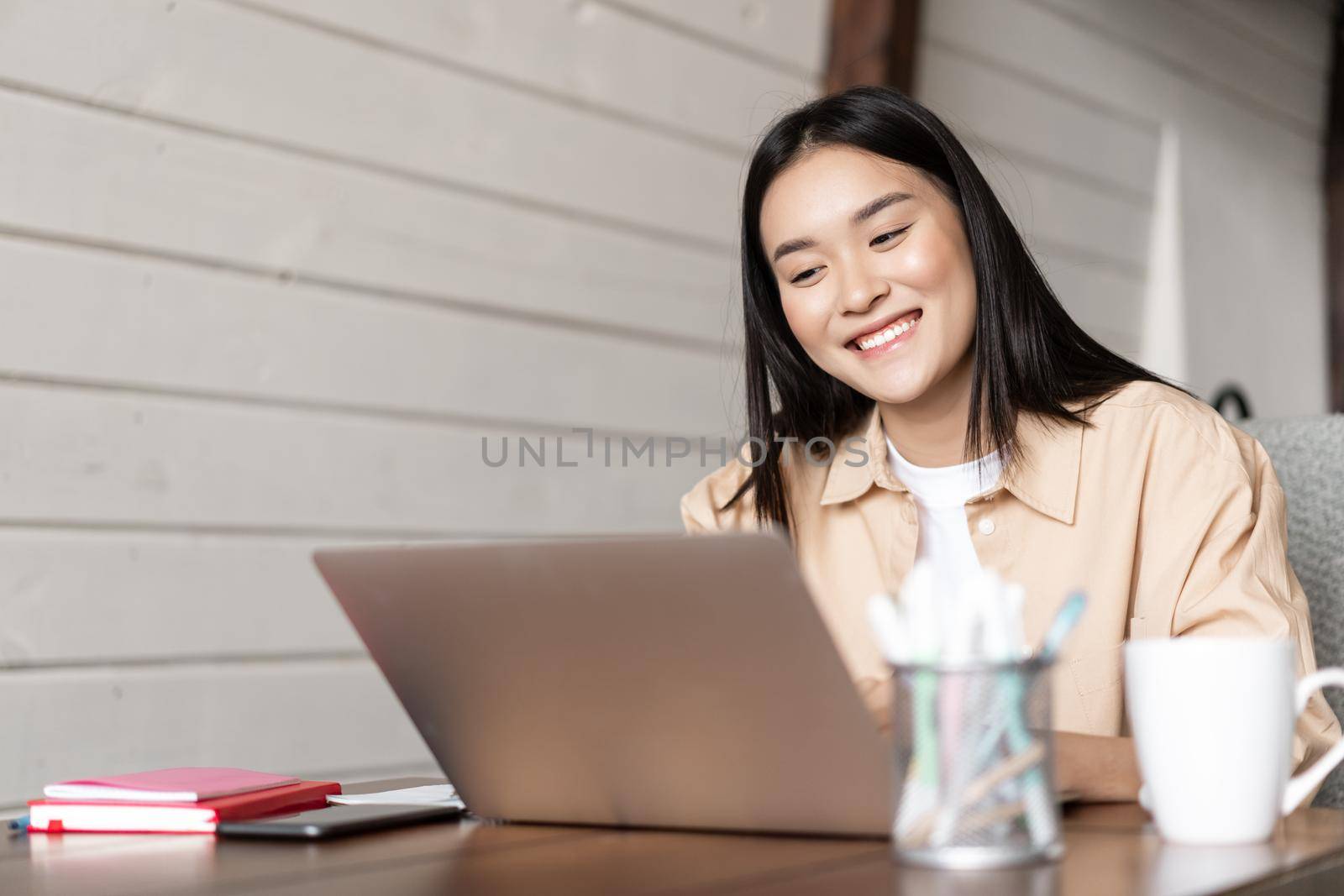 Smiling asian girl looks at laptop screen, works from home, listens webinar or attends online lecture, university classes on remote by Benzoix