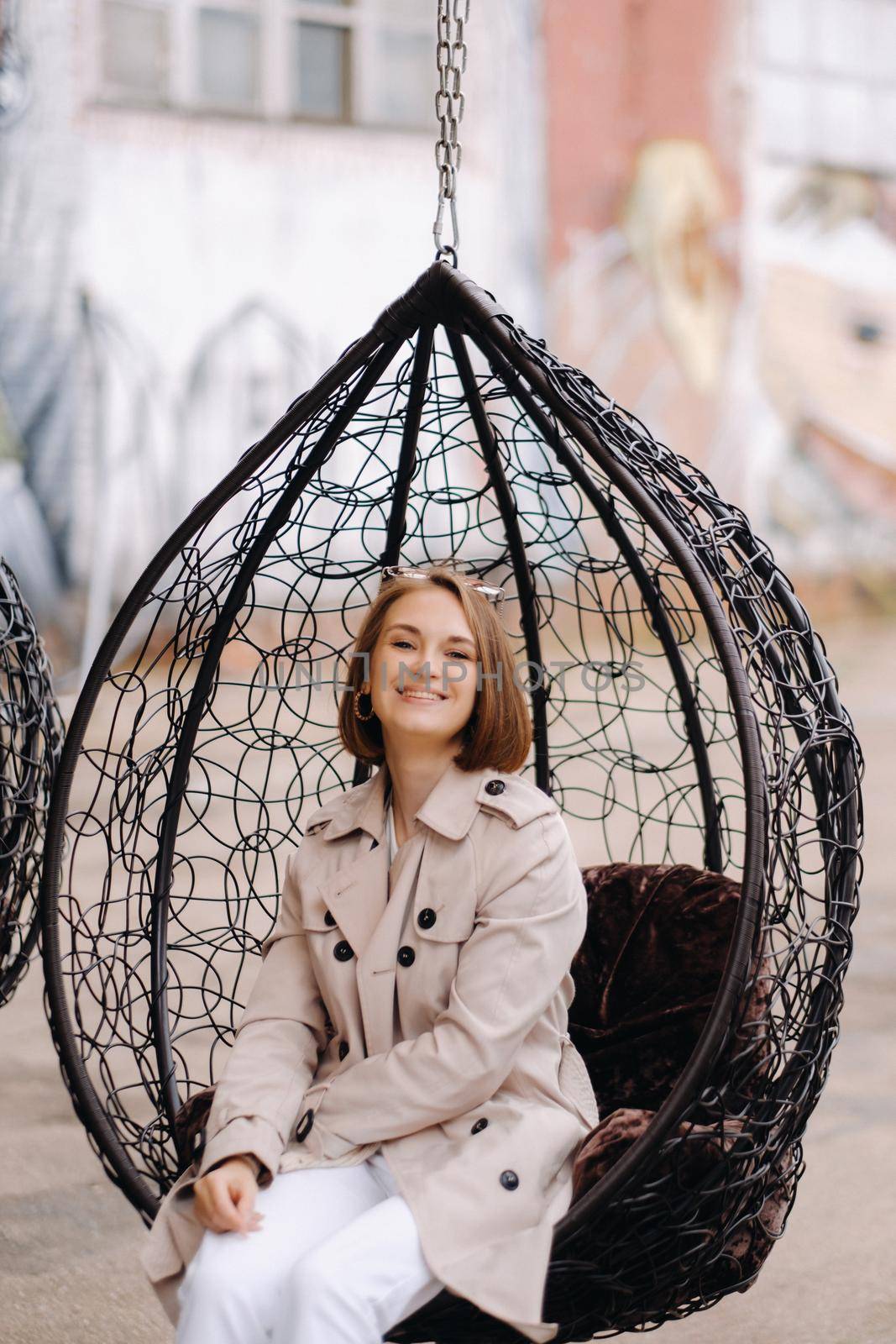 A happy stylish girl In a gray coat is sitting outside in an armchair by Lobachad