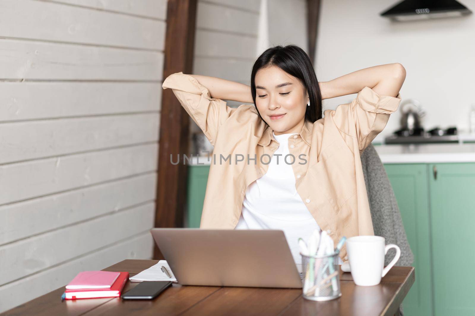 Image of smiling asian girl finish homework or work on laptop, looking pleased at result on computer screen, sitting at kitchen table as her workplace.