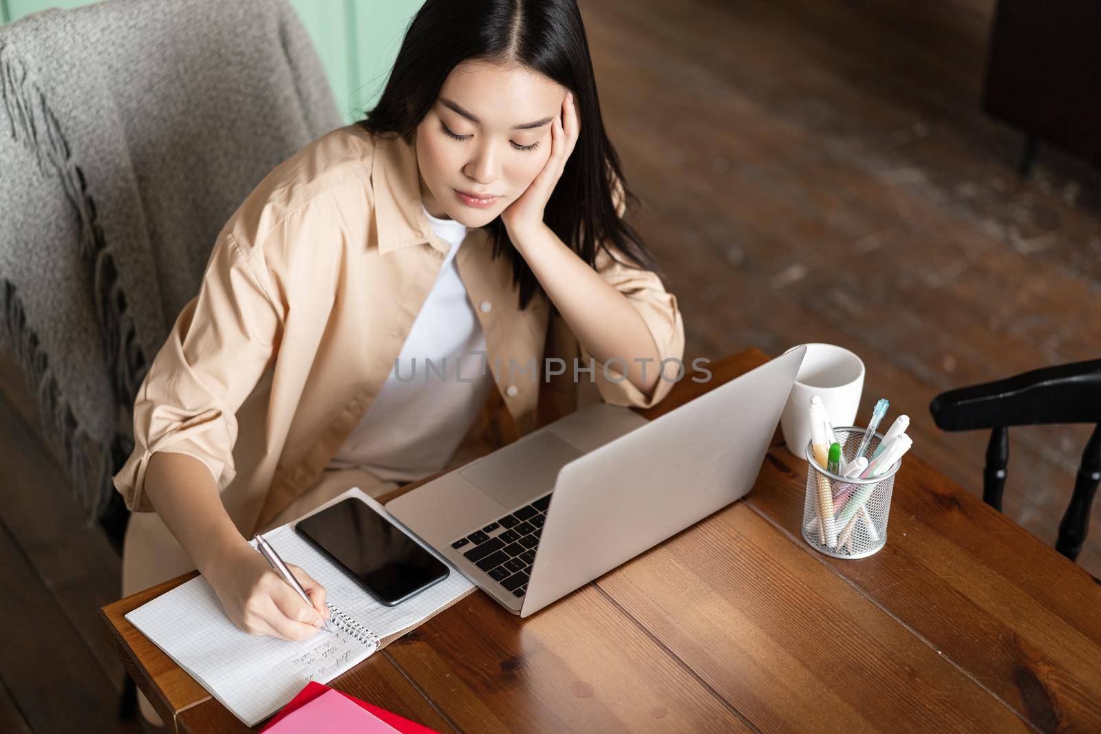 Focused asian girl doing homework, writing notes and sitting near laptop, attend webinar, online courses in internet.