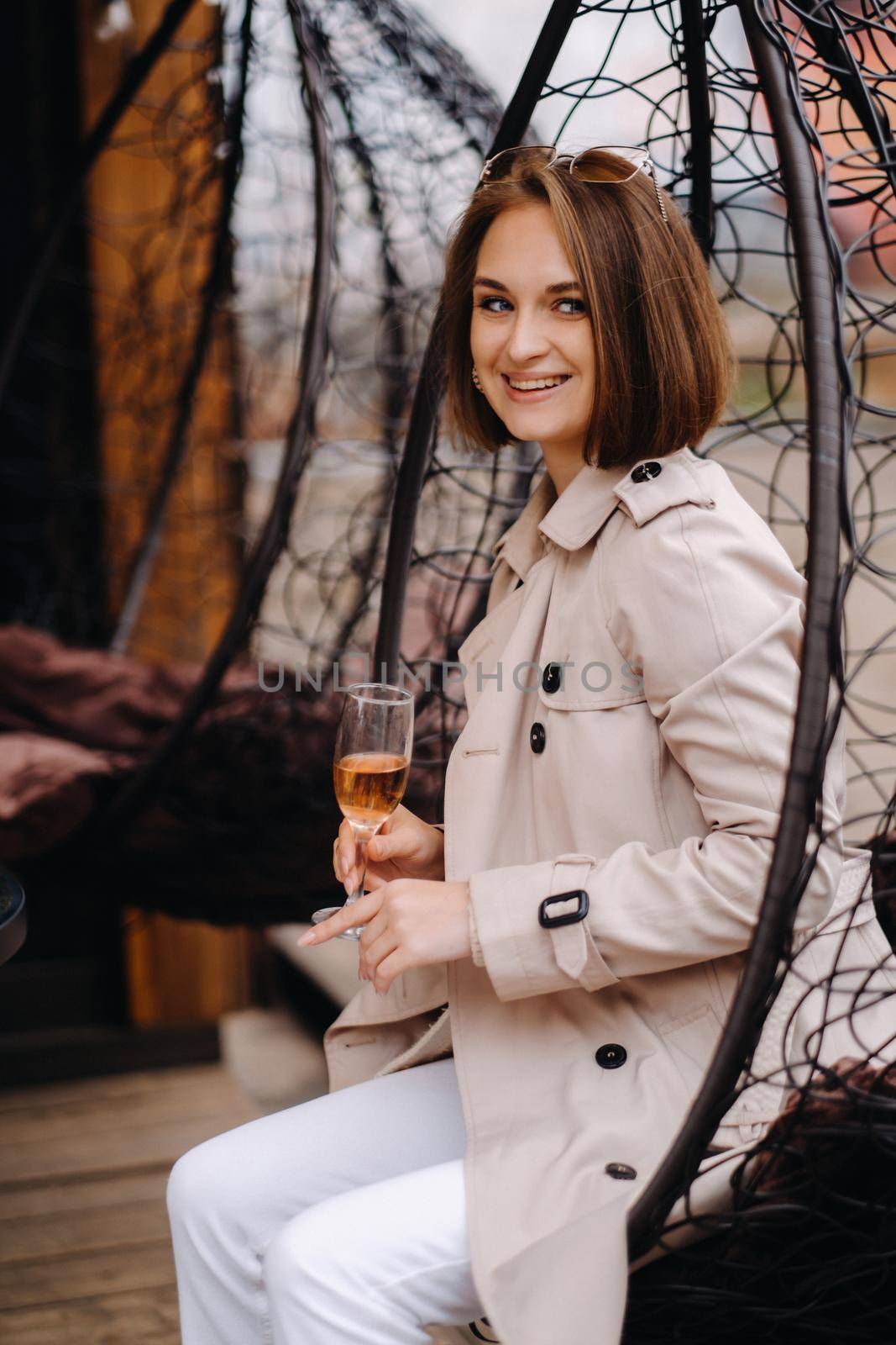 A happy stylish girl In a gray coat is sitting outside in an armchair and drinking a drink by Lobachad