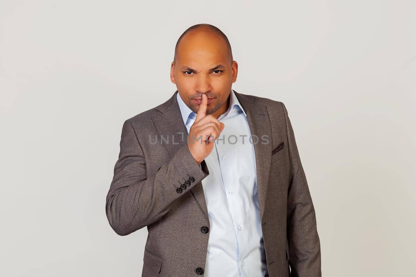 Portrait of a young African American guy businessman holding a finger on his lips, shows to close his mouth and stop making noise at the wrong moment. Standing on a gray background