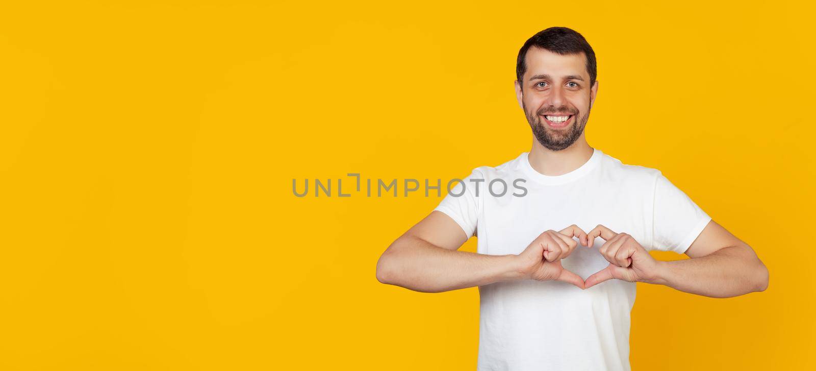 Handsome young man, a guy with a beard in a white T-shirt, smiles and makes a heart-shaped symbol with his finger, expresses love and a positive romantic feeling. Yellow background. by ViShark