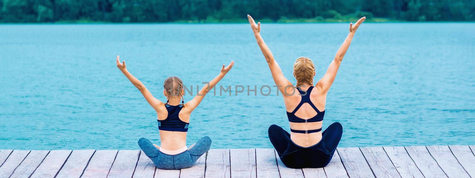 Mother and daughter doing yoga at the shore of the lake in summer.