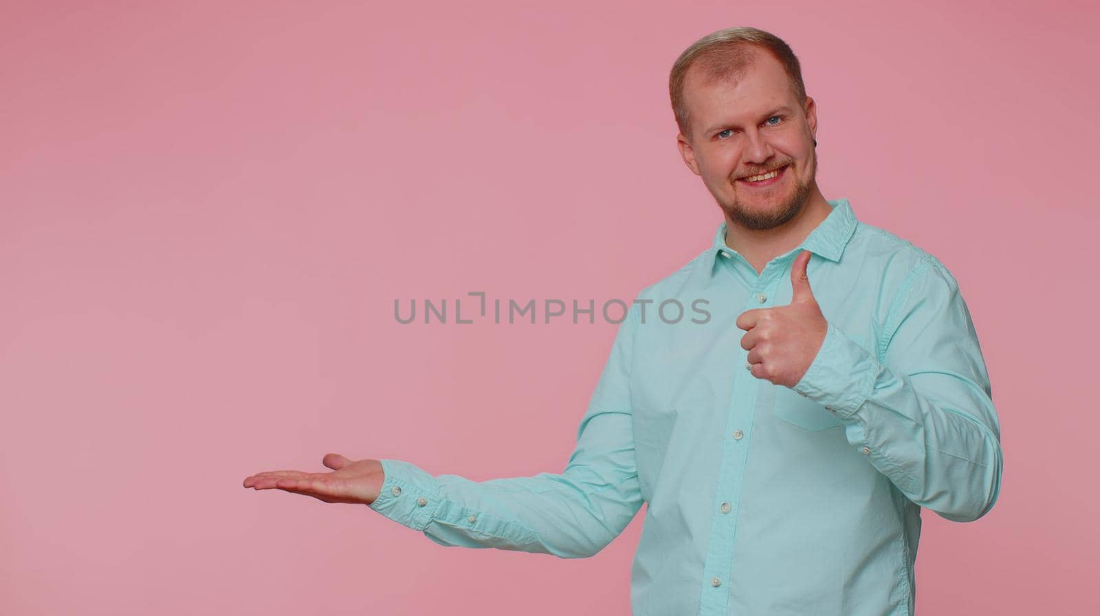 Handsome one bearded man showing thumbs up and pointing empty place, advertising area for commercial text, copy space for goods promotion. Young adult guy. Studio shot indoors on pink wall background