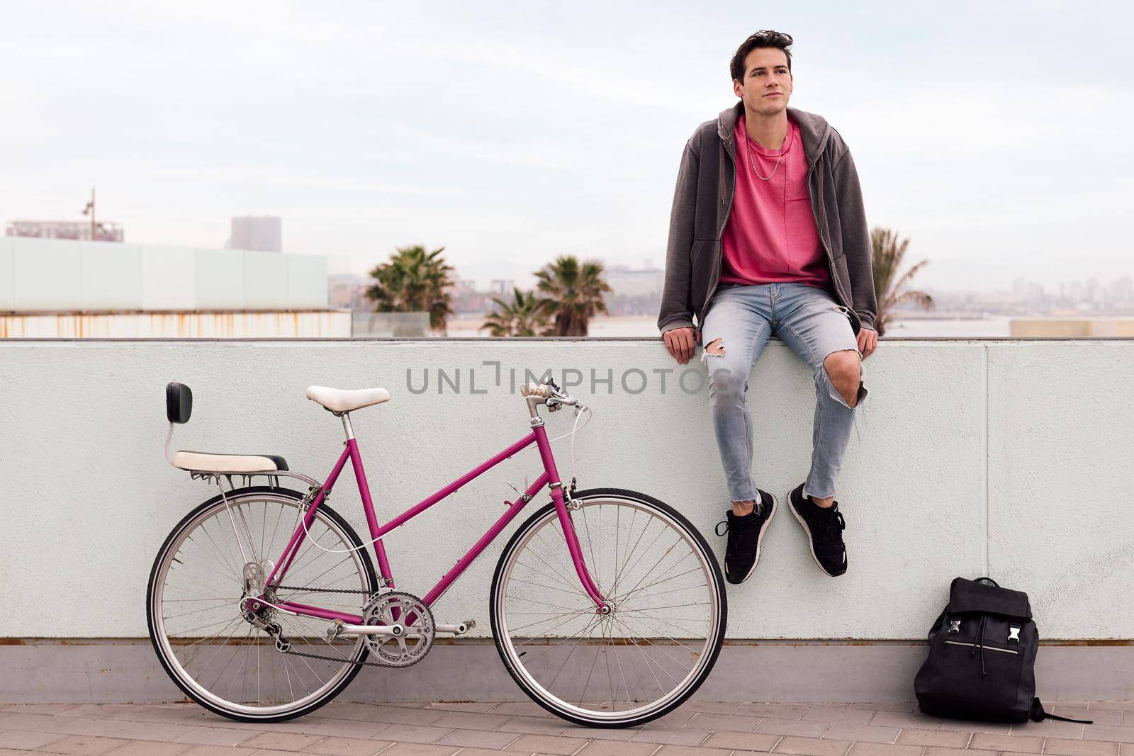 attractive young student sitting pensive with his vintage bike and backpack, concept of sustainable transportation and urban lifestyle, copy space for text