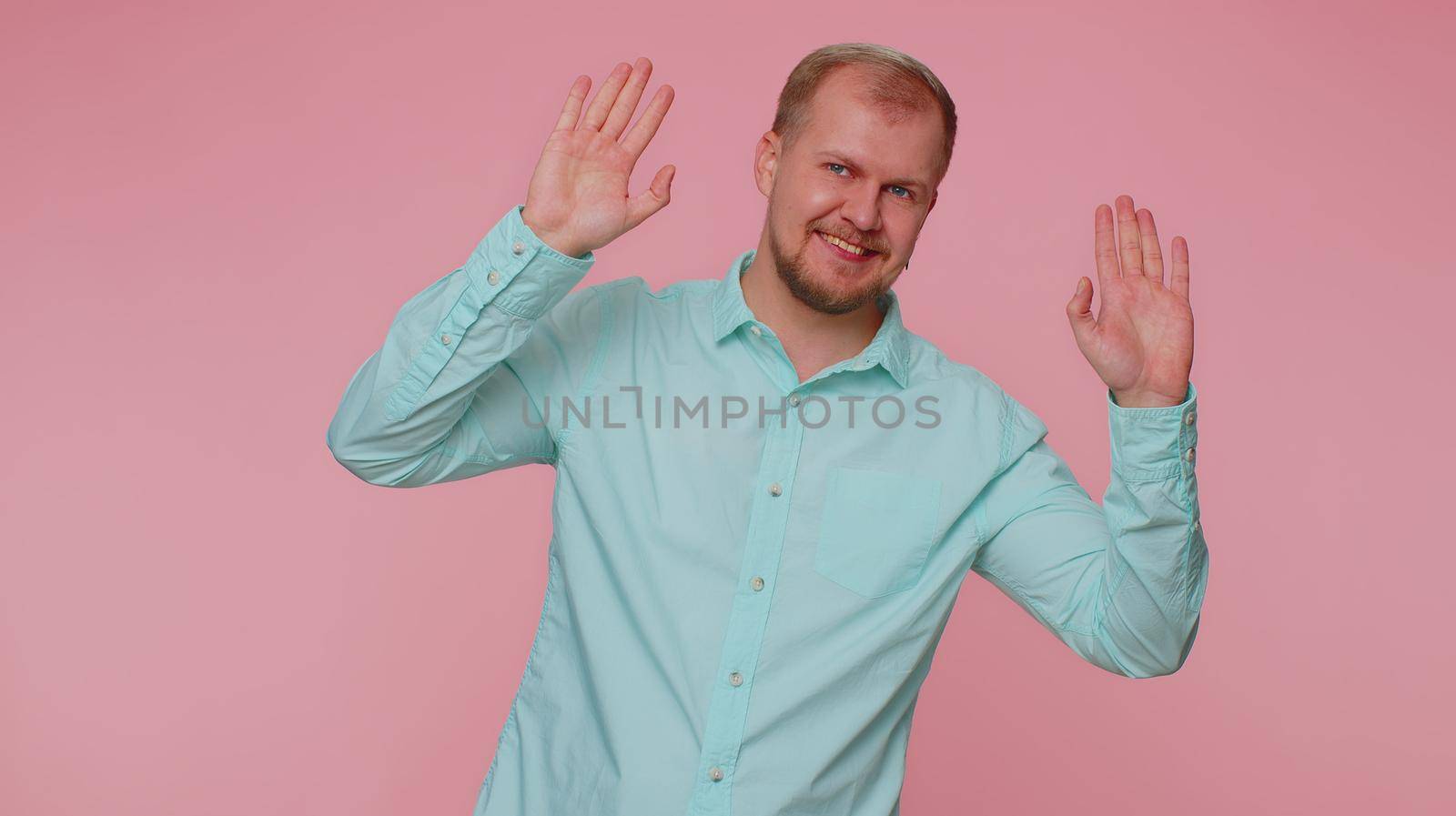 Handsome friendly man in shirt smiling friendly at camera and waving hands gesturing hello or goodbye, welcoming with hospitable expression. Young happy guy indoors isolated on pink wall background