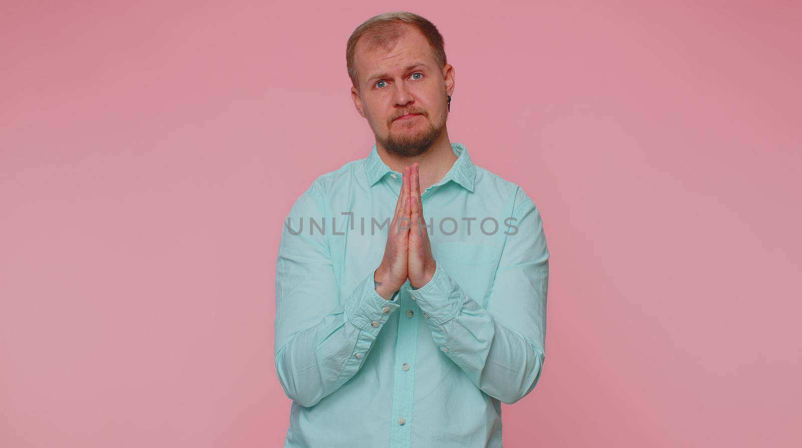 Please, God help. Sad worried man praying, looking upward, making wish asking with hopeful imploring expression begging apology, forgiveness. Young adult guy indoor isolated alone on pink background