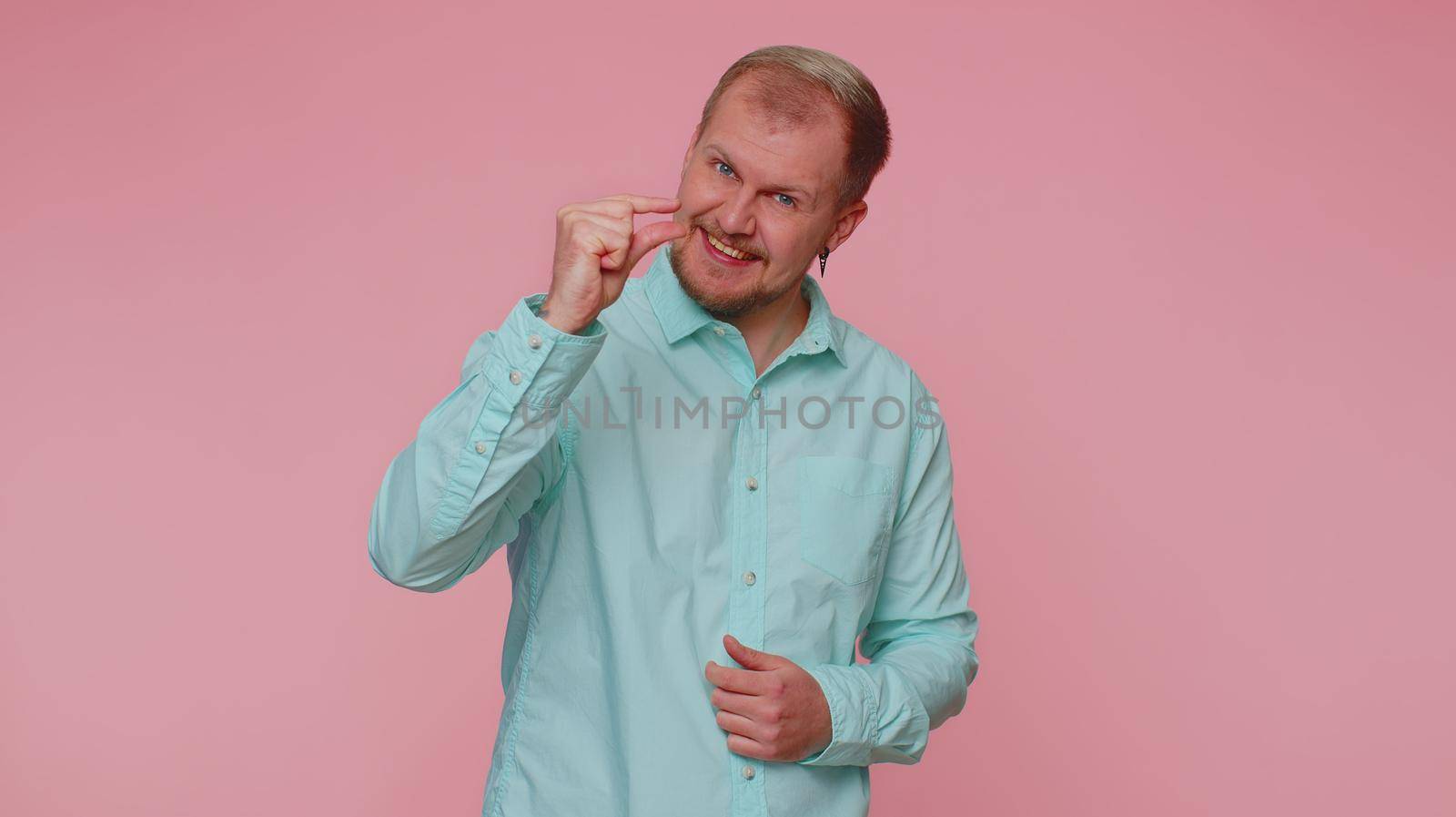Need some more, please give me. Man showing a little bit gesture with sceptic smile, showing minimum sign, measuring small size, asking for help. Young guy isolated alone on pink studio background