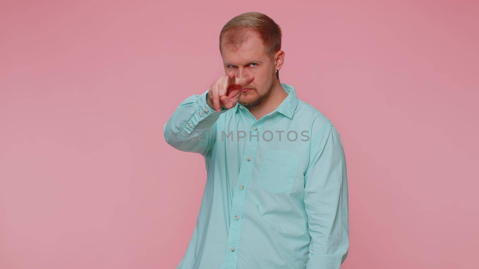 I am watching you. Man in blue casual shirt pointing at his eyes and camera, show I am watching you gesture, spying on someone. Young adult blonde guy boy isolated alone on pink studio wall background