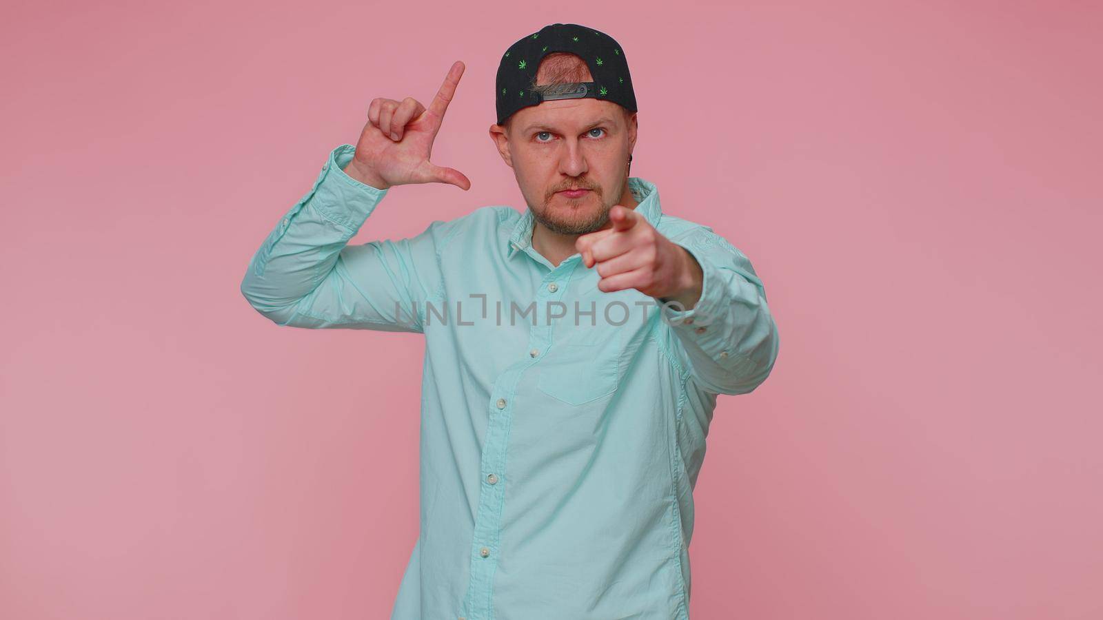 Man showing loser gesture and pointing on you, blaming accusing for unsuccess, expressing disrespect, mocking your failure, bullying abuse. Young guy isolated alone on pink studio wall background