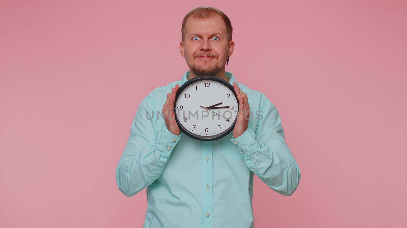 Joyful young man holding clock watch, hiding, checking time on clock, running late to work, being in delay, deadline on pink studio background. Serious guy looking at camera worrying to be punctual