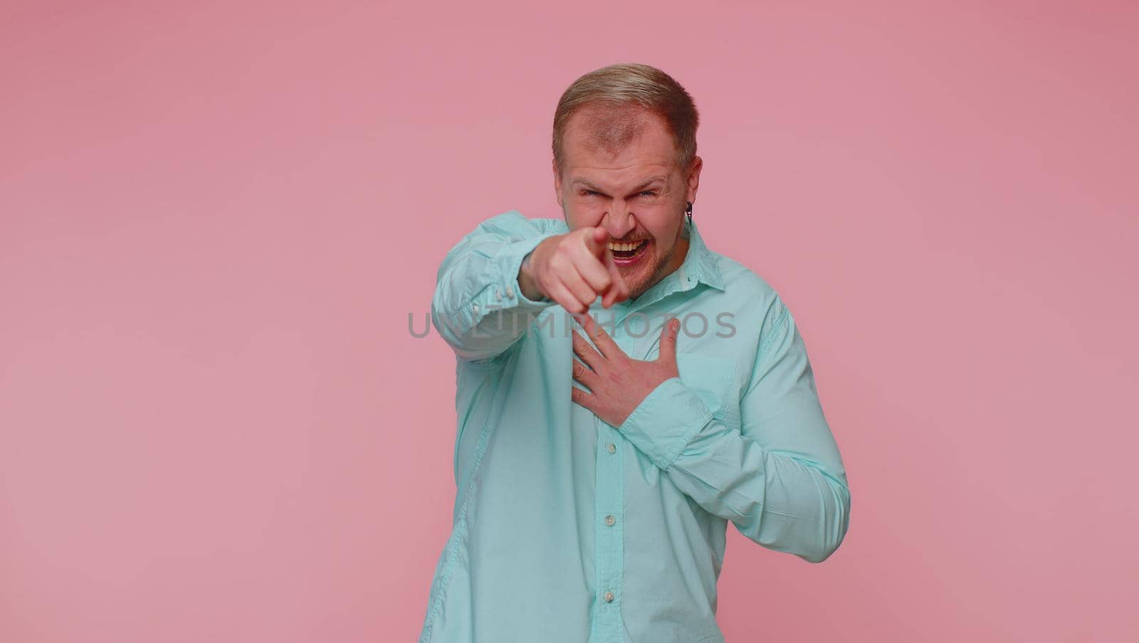 Amused man in casual blue shirt pointing finger to camera, laughing out loud, taunting making fun of ridiculous appearance, funny joke. Young guy boy posing alone on pink studio wall background