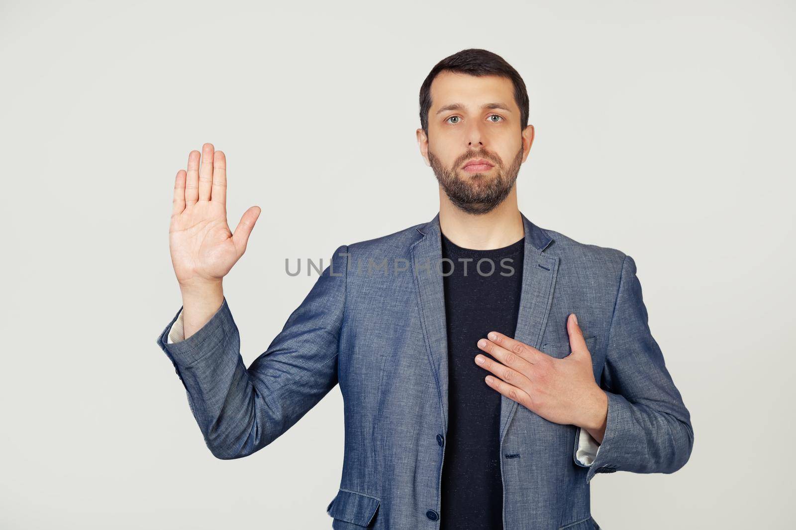 Young businessman man with a beard in a jacket, swears, putting his hand on his chest and open palm, making an oath of promise of loyalty. Portrait of a man on a gray background.
