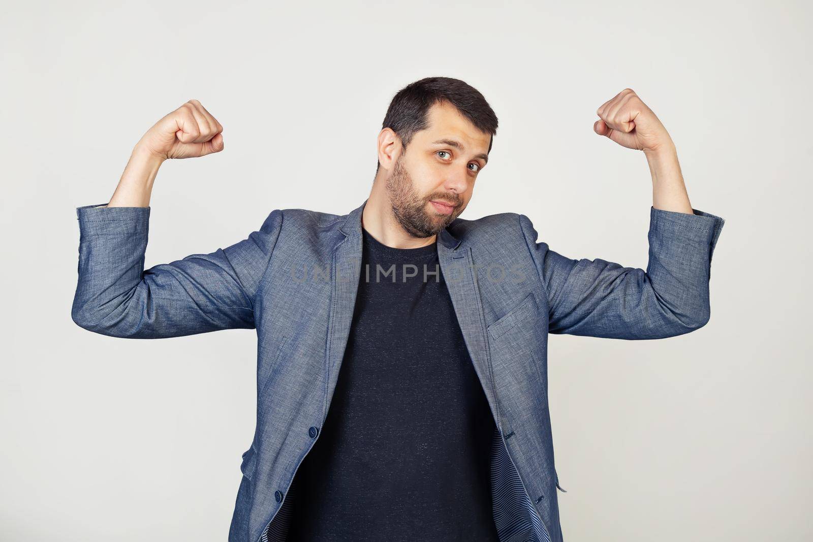 Young smiling businessman man with beard in jacket showing arm muscles, smiling proudly. Fitness concept. Portrait of a man on a gray background.