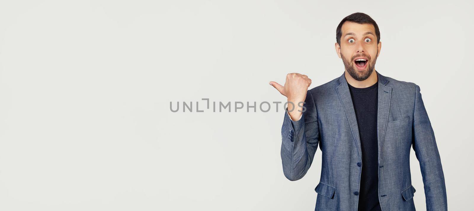 Young businessman man with a beard in a jacket, surprised, pointing finger to the side, with open mouth amazed expression. Portrait of a man on a gray background.