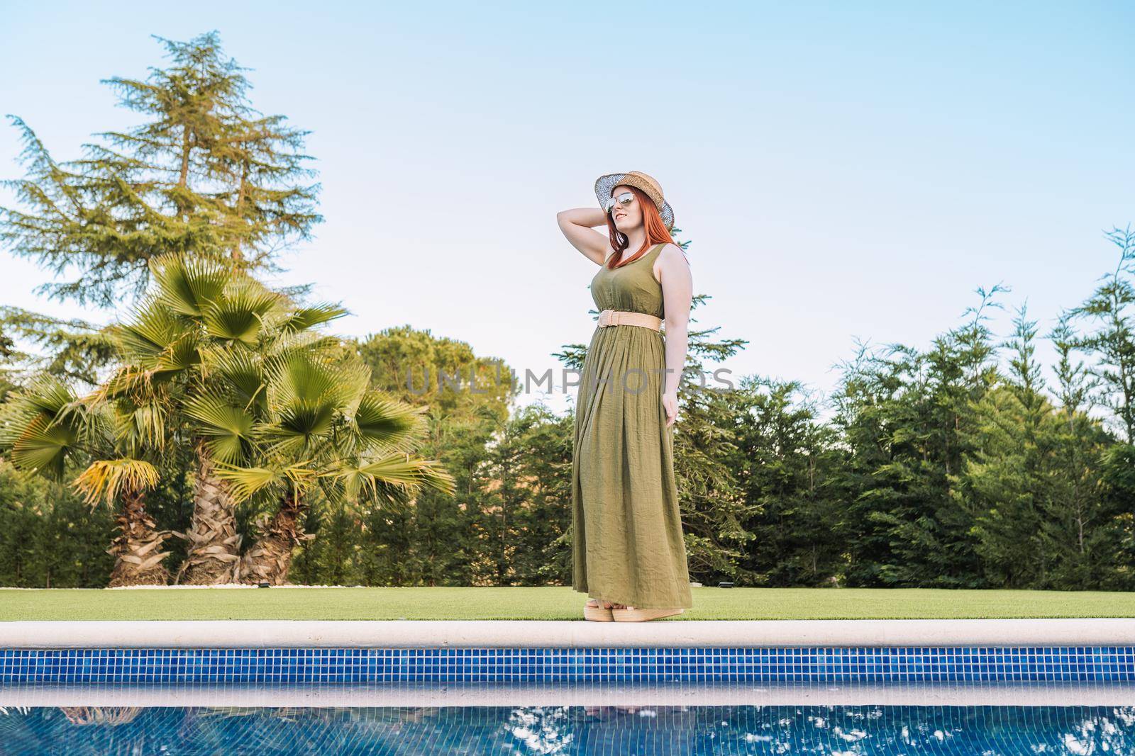 young woman traveller, standing sunbathing in front of the swimming pool of her holiday hotel. woman on a leisure trip in the resort garden. by CatPhotography