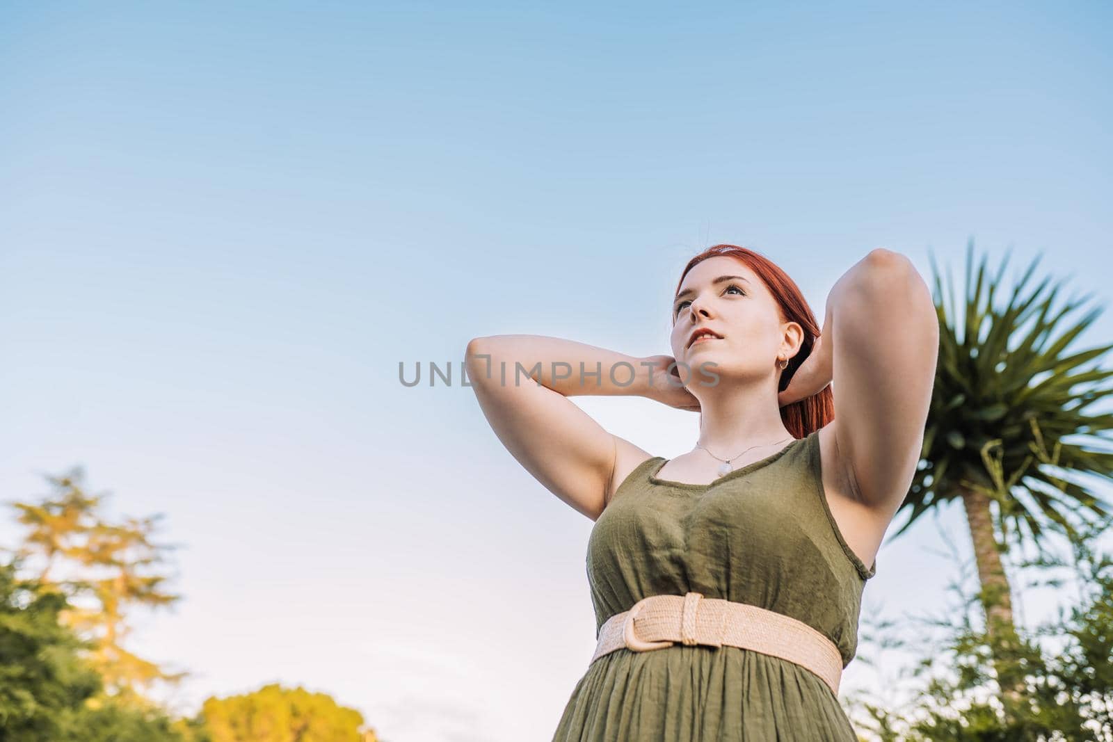 young adventurous girl, relaxing on her summer holiday. woman bathed in sunlight holding up her hair. concept trip and leisure. woman in green summer dress, straw hat, outdoors with natural light and plants in background.