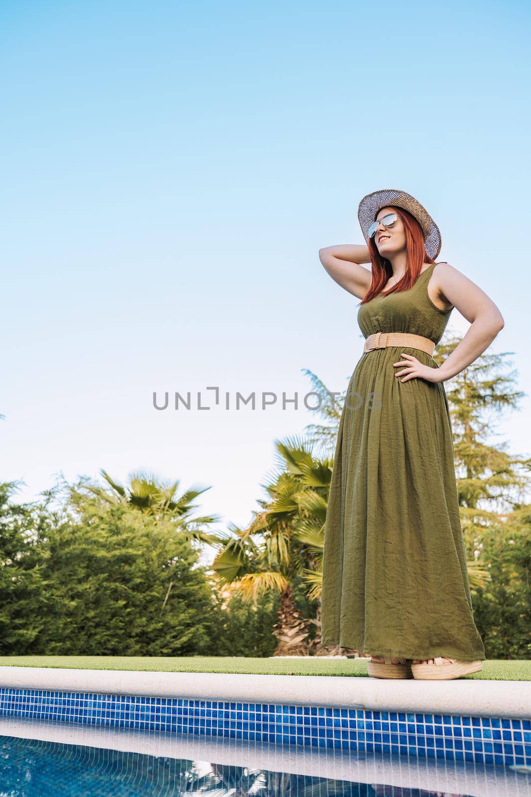 adventurous young woman, standing sunbathing in front of the swimming pool of her holiday hotel. woman on recreational trip. Outside, dim sunlight, grass floor with white tiles. vertical. copy space.