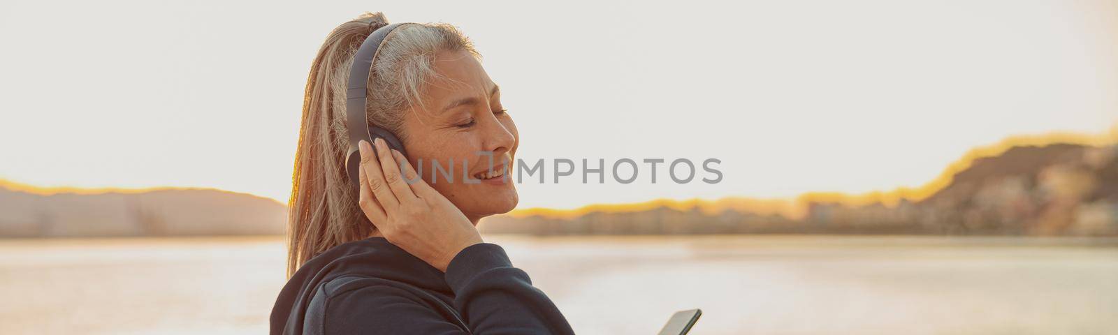 Portrait of calm mature woman standing on the seashore in the evening and listening to music with closed eyes