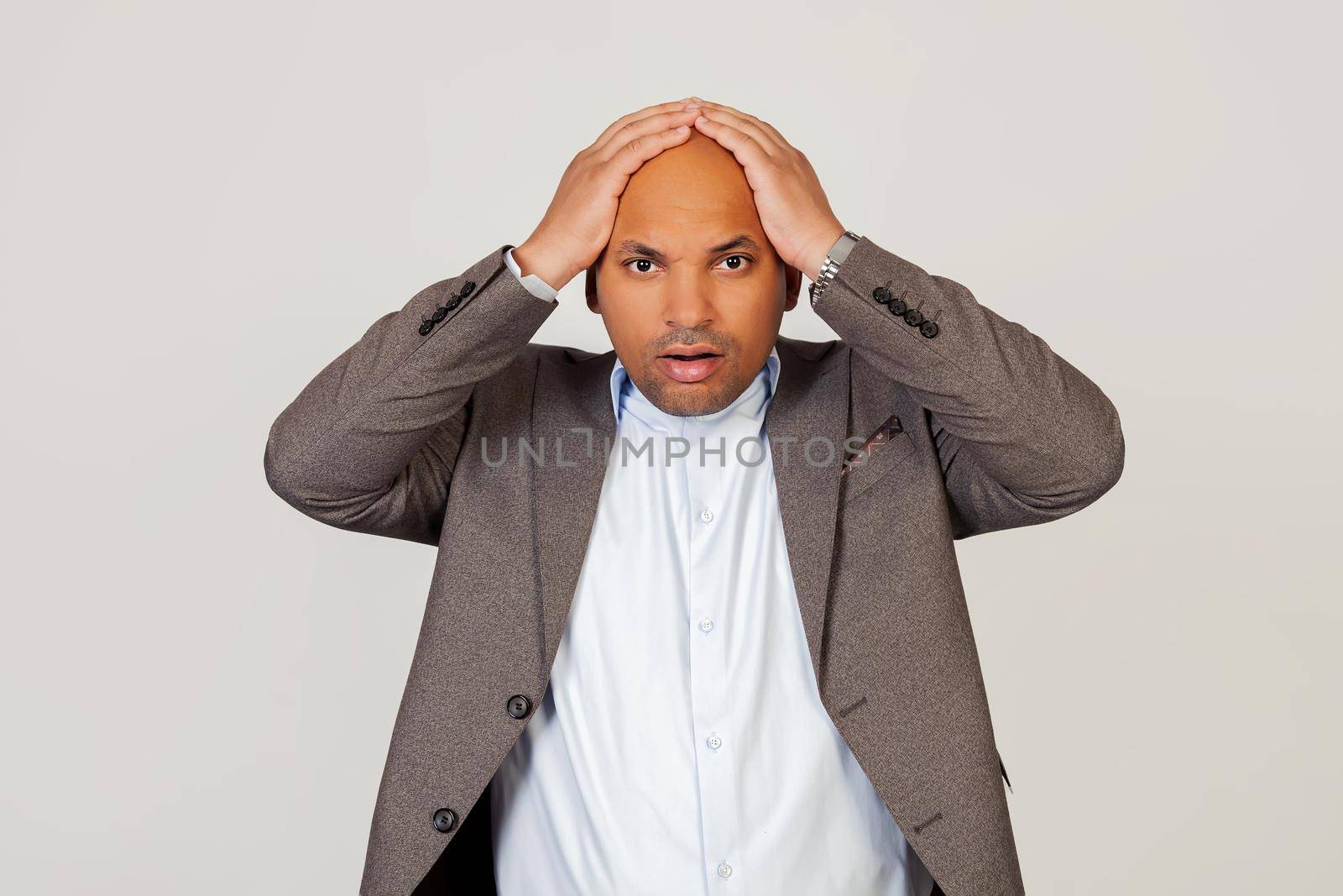 Surprised young male African American businessman holding his hands behind his head, absolutely shocked by the news. standing on a gray background. Human face expression concept