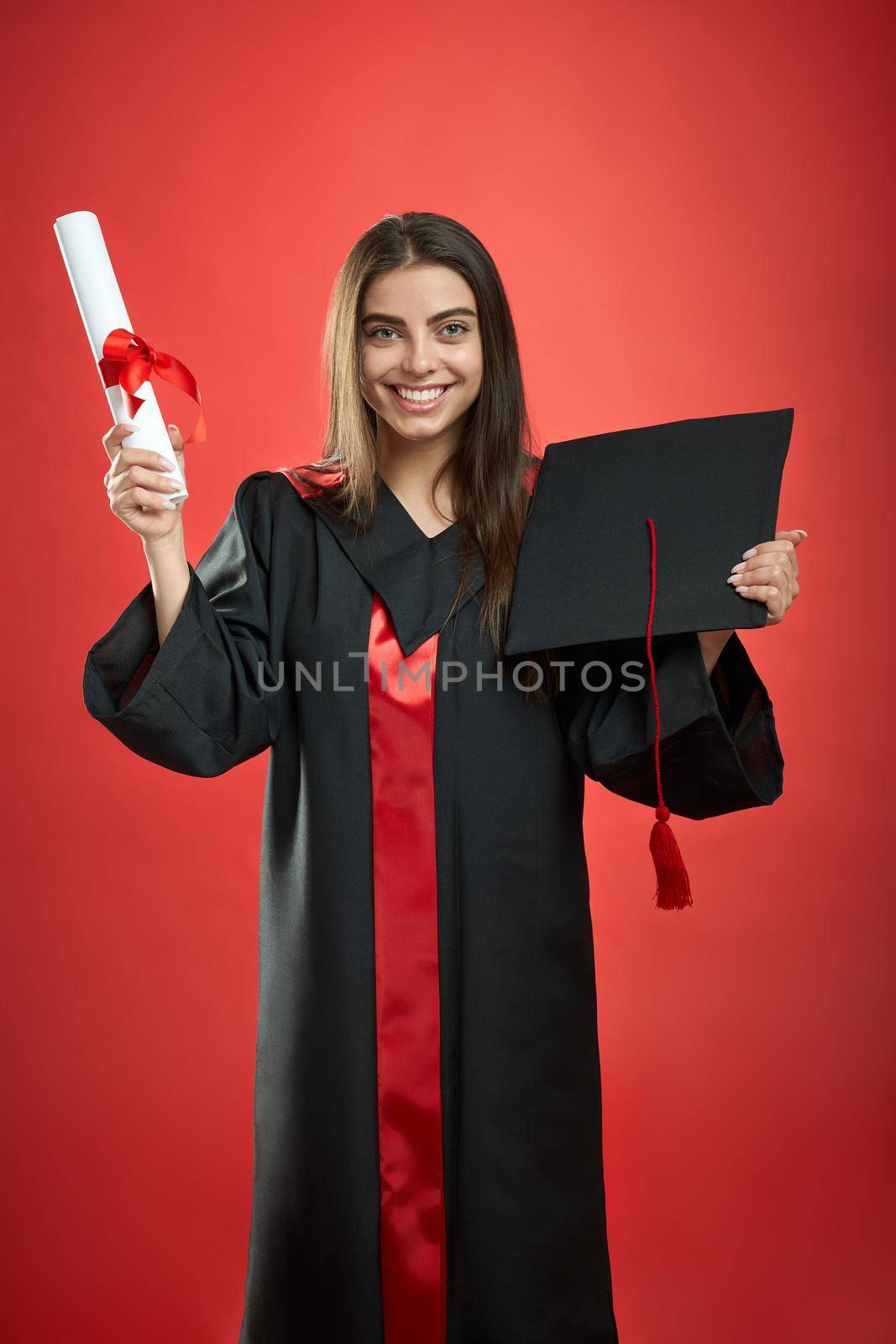 Front view of brunette girl graduating from college, university, high school. Pretty girl looking at camera, smiling, holding diploma, mortarboard. Isolated on red background,