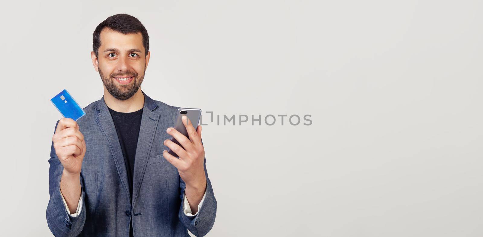 Young businessman man with a beard in a jacket, Handsome man using a credit card to pay online using a smartphone. Portrait of a man on a gray background.