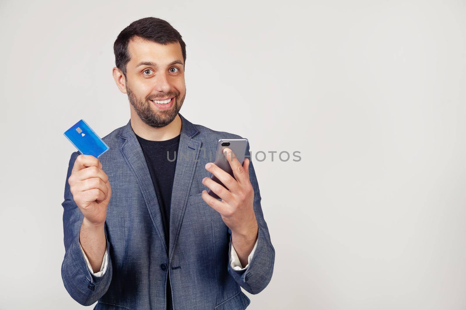 Young businessman man with a beard in a jacket, Handsome man using a credit card to pay online using a smartphone. Portrait of a man on a gray background.