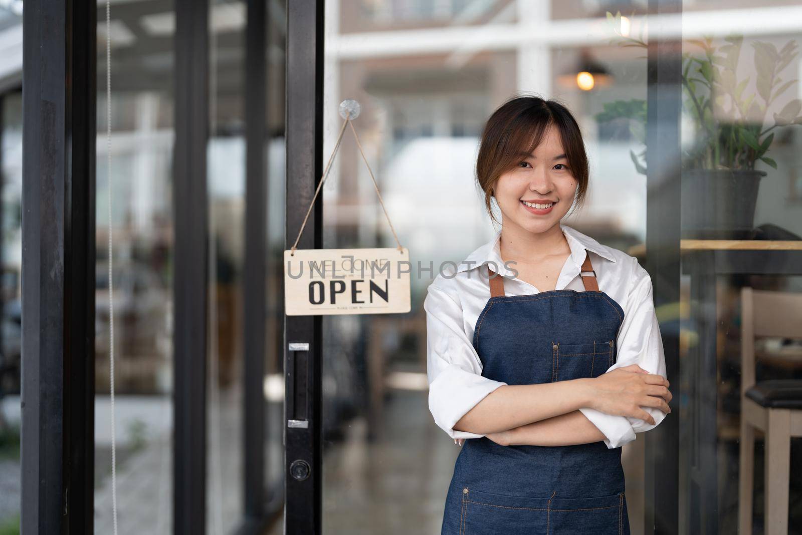 Beautiful asian young barista woman in apron holding tablet and standing in front of the door of cafe with open sign board. Business owner startup concept.