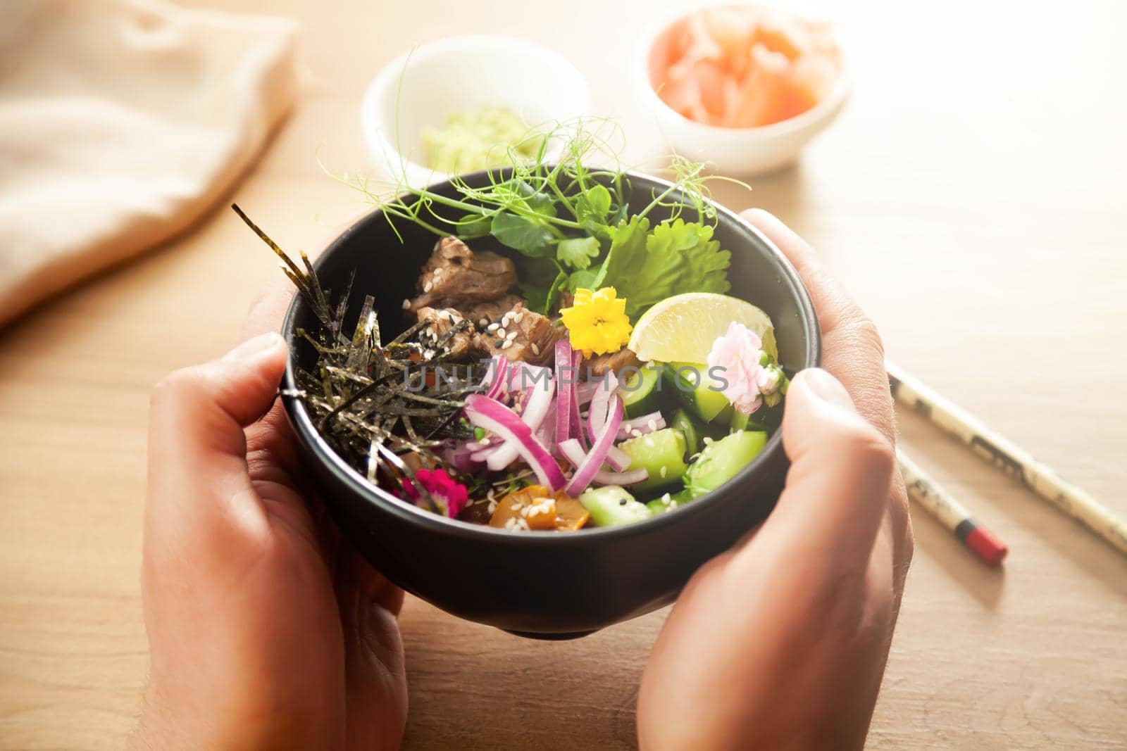 A man holds a Poke salad with beef in a bowl in his hands. Ingredients Beef, Nameko mushrooms, cherry tomatoes, rice, cucumber, red onion, sesame seeds, cilantro, lime. Asian salad concept.