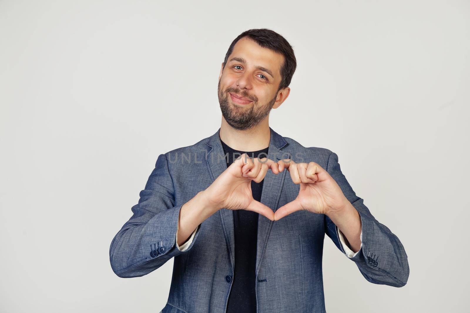Young smiling businessman man with a beard in a jacket, smiling and making a heart shaped symbol with a finger, expressing love and positive romantic feeling. Portrait of a man on a gray background.