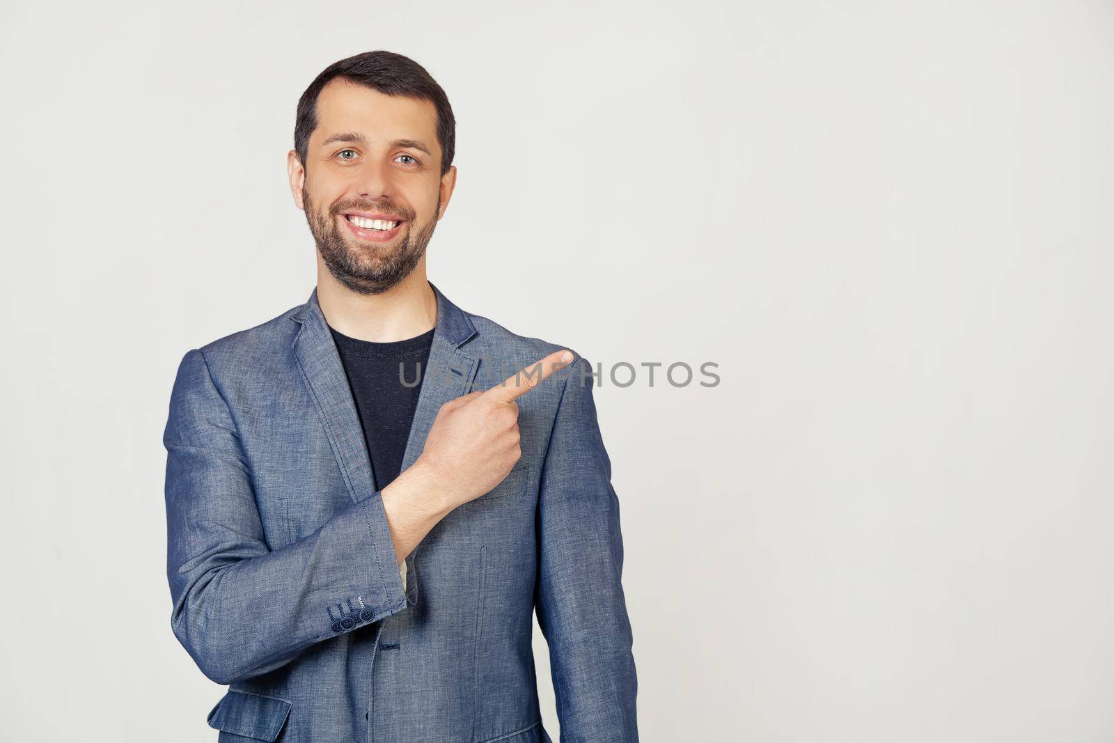 Young businessman man with a beard in a jacket, cheerful, with a smile on his face, pointing with his hand and finger to the side with a happy and natural expression. Portrait of a man on a gray background.