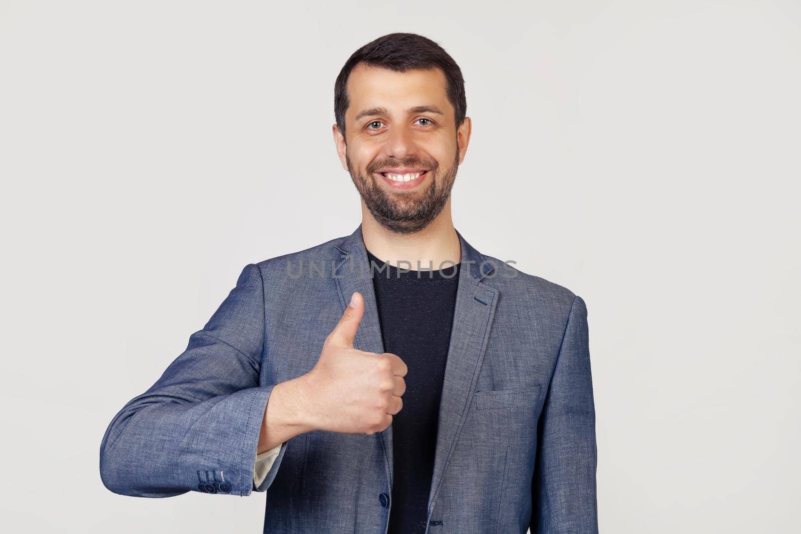 Young businessman man with a beard in a jacket, with a happy smile, shows his thumb to the camera. All is well, it shows success. Portrait of a man on a gray background