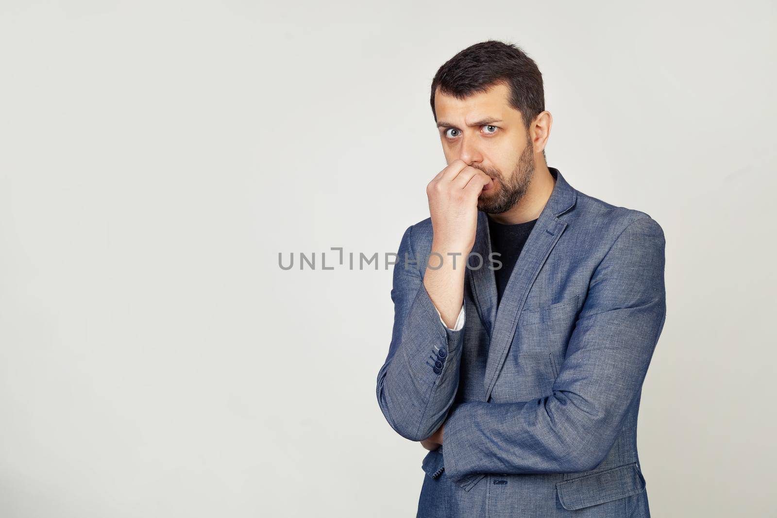 Young businessman with a smile, a man with a beard in a jacket, looks tense and nervous with hands on his lips, biting his nails. Anxiety problem. Portrait of a man on a gray background.