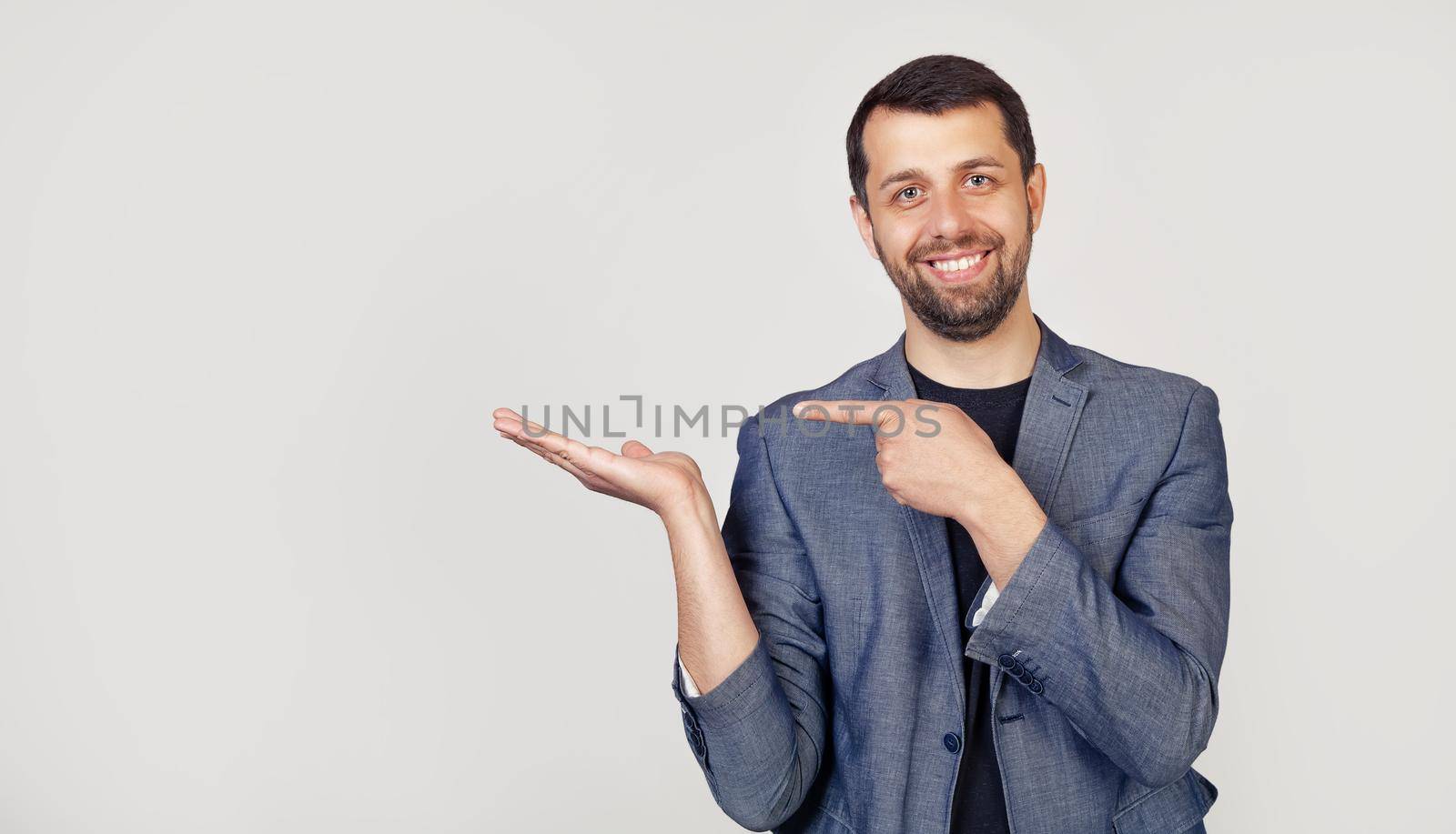 Young businessman man with a beard in a jacket, amazed and smiling at the camera, showing with his hand and pointing his finger. Portrait of a man on a gray background.